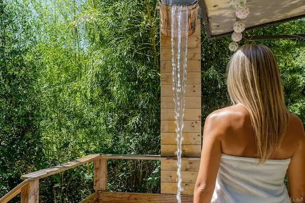 Femme de dos face à une douche extérieure en cascade, entourée de bambous, après une séance de sauna.