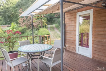 Terrasse en bois du lodge avec table ronde, chaises, pergola et vue sur le jardin arboré.