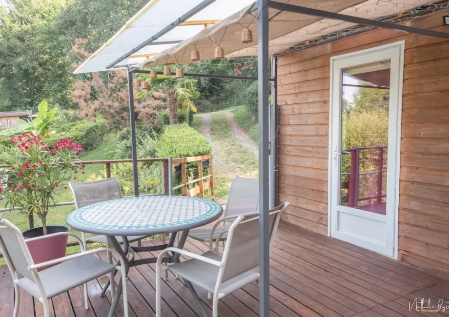 Terrasse en bois du lodge avec table ronde, chaises, pergola et vue sur le jardin arboré.