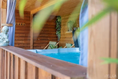 Jacuzzi bleu en bois avec eau bouillonnante, fauteuils de détente et décoration végétale dans une alcôve couverte.