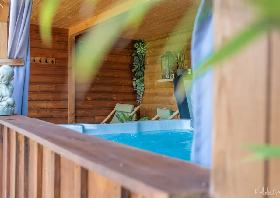 Jacuzzi bleu en bois avec eau bouillonnante, fauteuils de détente et décoration végétale dans une alcôve couverte.